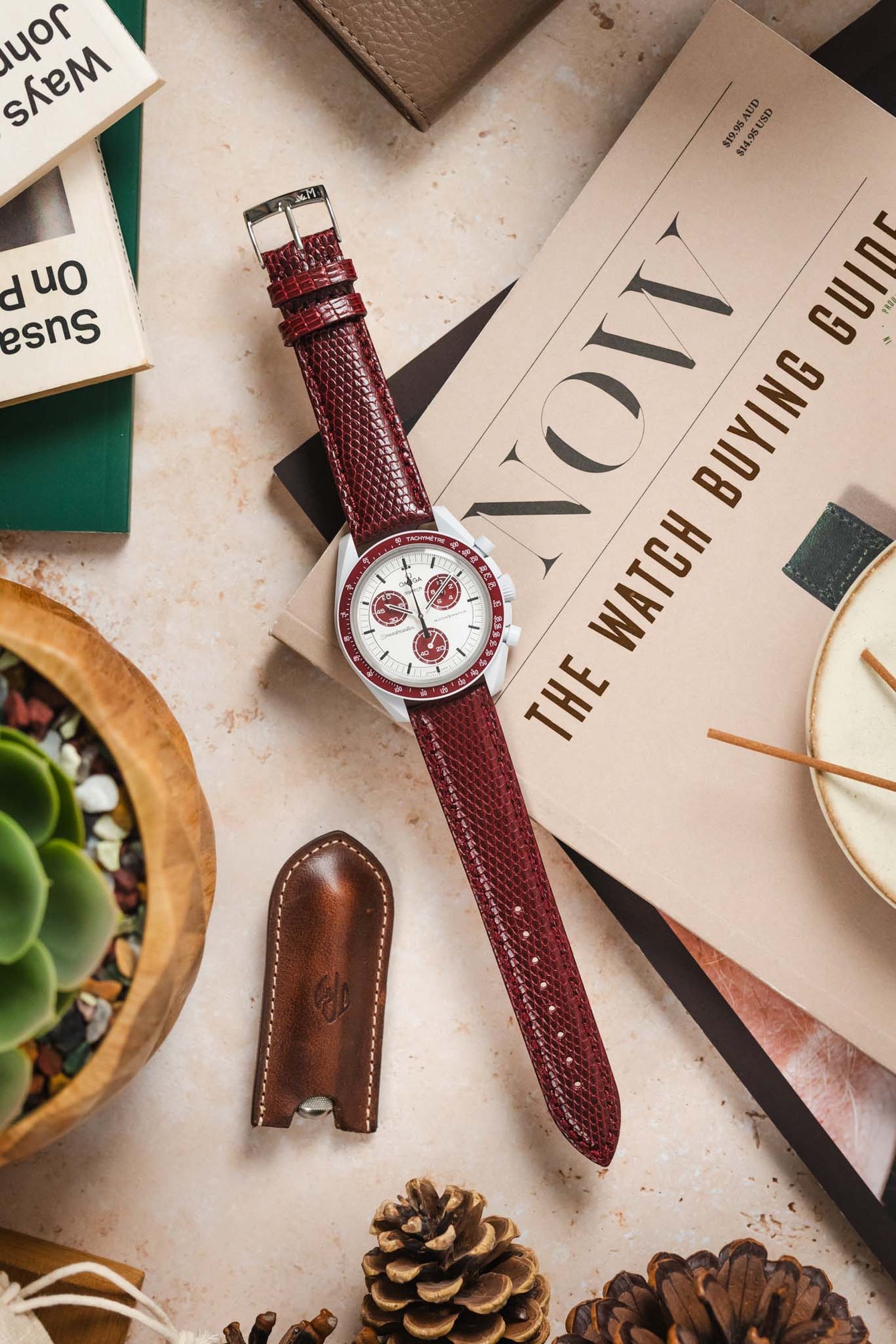 
A top-down flat lay composition showcasing a white Omega x Swatch MoonSwatch "Alaska Project" or "Arctic" watch with red accents and a long, textured deep burgundy watch strap with a silver buckle. The watch is centrally placed, resting on a magazine titled "NOW THE WATCH BUYING GUIDE". It is surrounded by various objects on a light, textured surface, including pinecones, a brown leather watch tool holder, a small potted succulent plant, and parts of books.