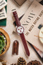 
A top-down flat lay composition showcasing a white Omega x Swatch MoonSwatch "Alaska Project" or "Arctic" watch with red accents and a long, textured deep burgundy watch strap with a silver buckle. The watch is centrally placed, resting on a magazine titled "NOW THE WATCH BUYING GUIDE". It is surrounded by various objects on a light, textured surface, including pinecones, a brown leather watch tool holder, a small potted succulent plant, and parts of books.