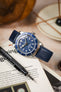 A top-down, slightly angled shot of a silver Tudor Black Bay 58 watch with a blue dial and bezel, fitted with a dark blue lizard-patterned strap. The watch is resting on an open book or magazine. A black watch tool is positioned next to it. The background is blurred with warm, neutral tones, showing a decorative plate and part of a wooden object.