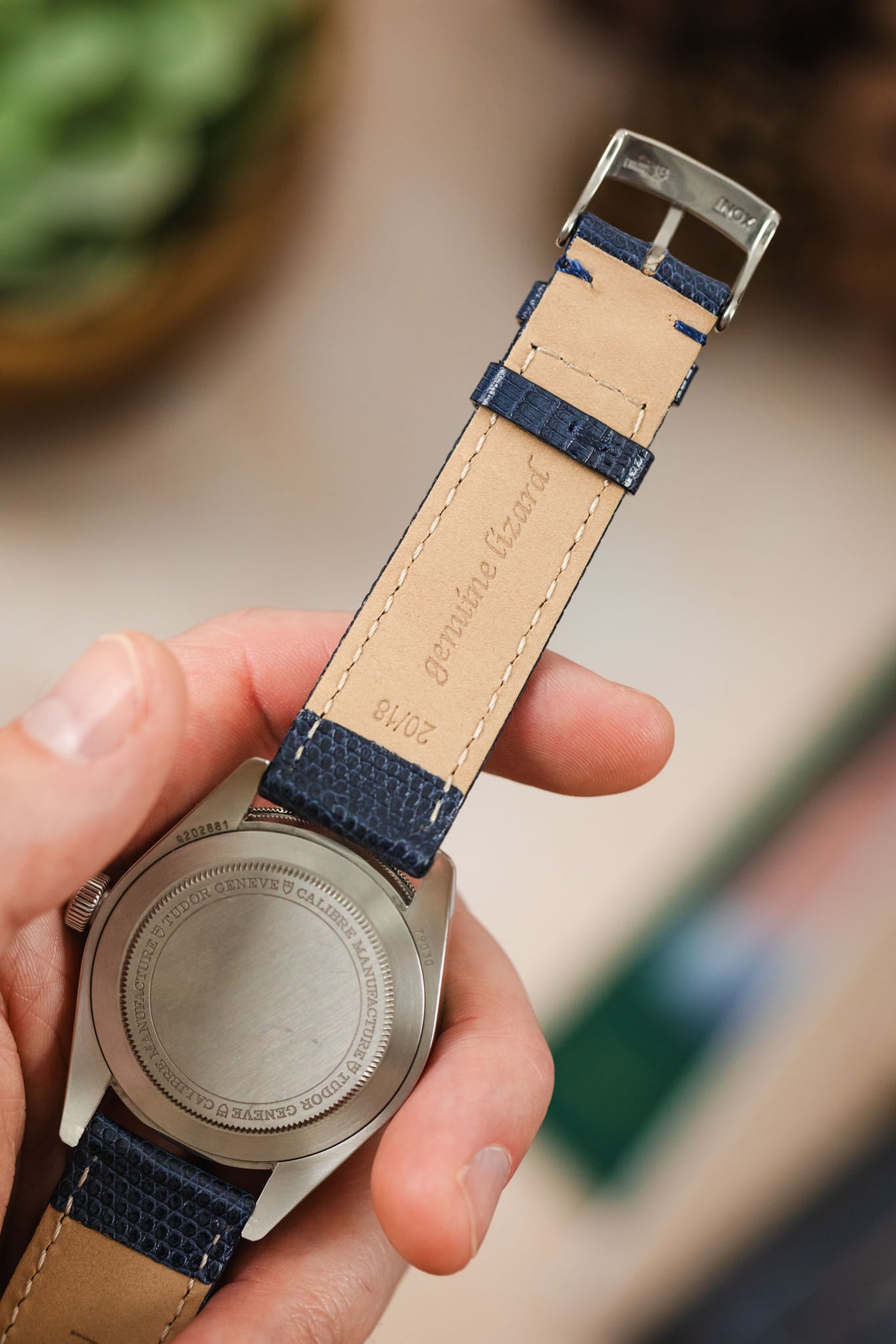 A hand holds the back of a silver Tudor Black Bay 58 watch, revealing its case back and a dark blue lizard-patterned watch strap. The light brown underside of the strap, with the text "Genuine Lizard" and "20/18" stamped on it, is clearly visible. The background is blurred with light, warm tones and hints of green.