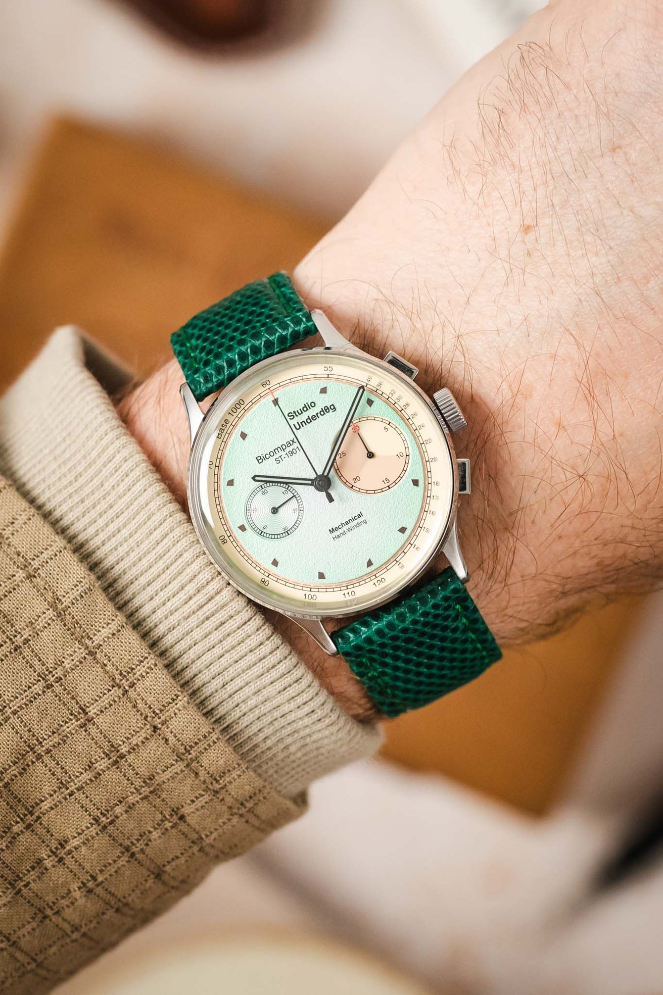 
A close-up shot of a wrist wearing a silver Studio Underd0g watch with a mint green and salmon-colored bicompax dial, paired with a vibrant green lizard-patterned watch strap. The arm has light skin and is partially covered by a beige, textured sleeve. The background is softly blurred with light, neutral tones.