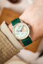 
A close-up shot of a wrist wearing a silver Studio Underd0g watch with a mint green and salmon-colored bicompax dial, paired with a vibrant green lizard-patterned watch strap. The arm has light skin and is partially covered by a beige, textured sleeve. The background is softly blurred with light, neutral tones.