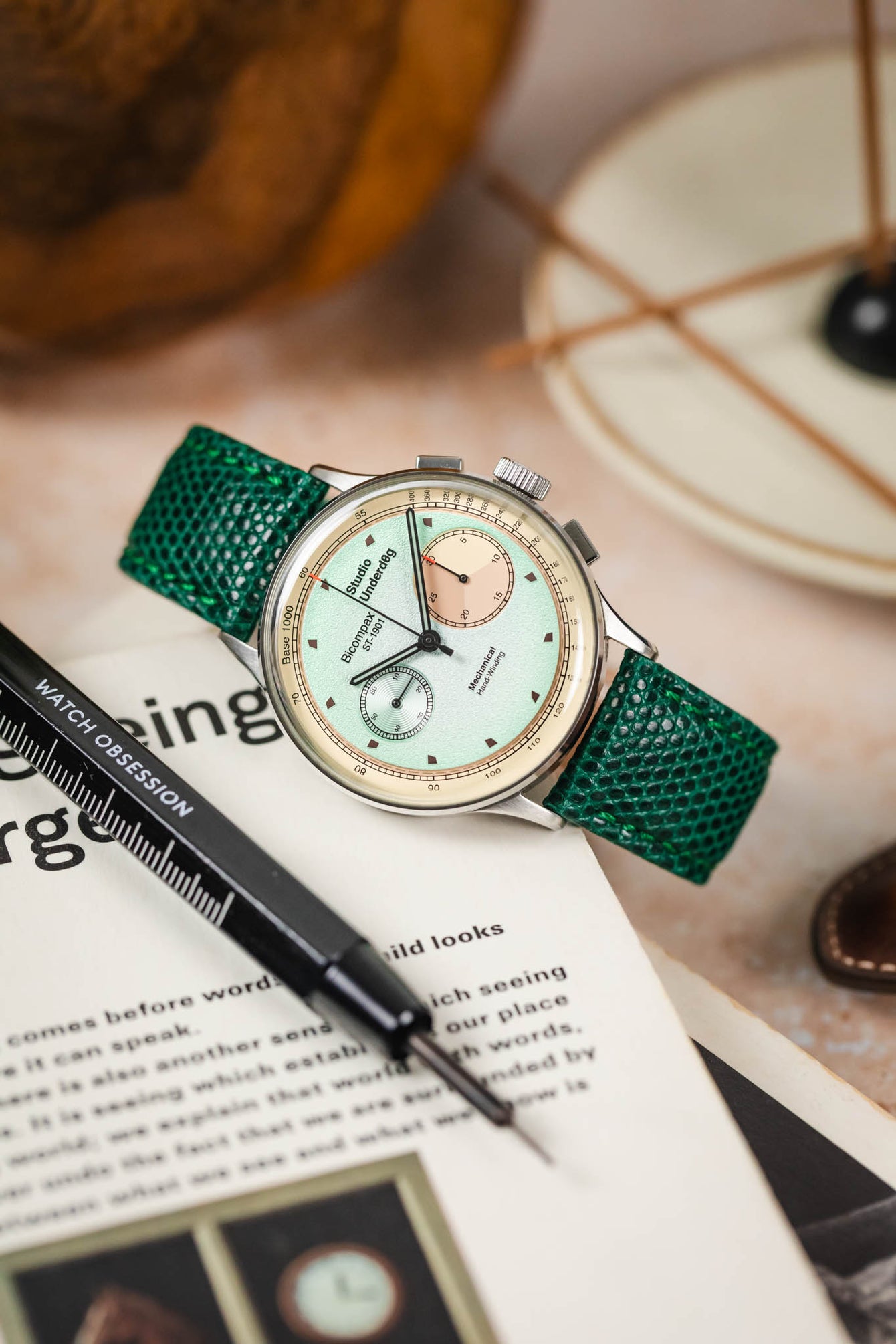 A top-down, slightly angled shot of a silver Studio Underd0g watch with a mint green and salmon-colored bicompax dial, fitted with a vibrant green lizard-patterned strap. The watch is resting on an open book or magazine with text, and a black watch tool is positioned next to it. The background is blurred with warm, neutral tones, showing a decorative plate and part of a wooden object.