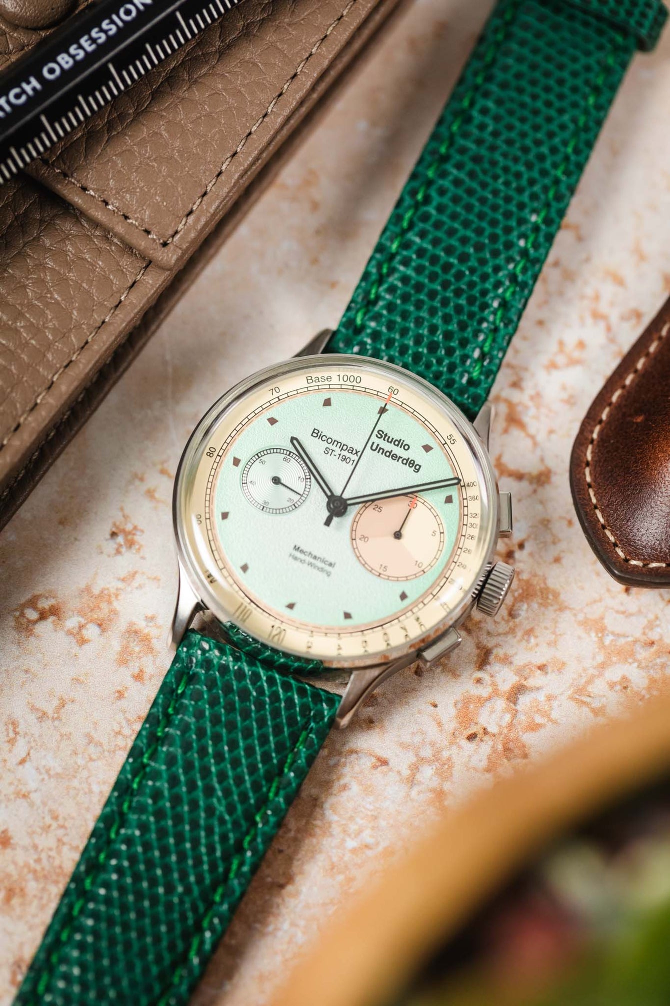 A close-up, angled shot focusing on a silver Studio Underd0g watch with a mint green and salmon-colored bicompax dial, fitted with a vibrant green watch strap featuring a lizard-skin texture. The watch is resting on a light, textured surface, with parts of a light brown leather watch pouch and a dark brown leather item blurred in the background.