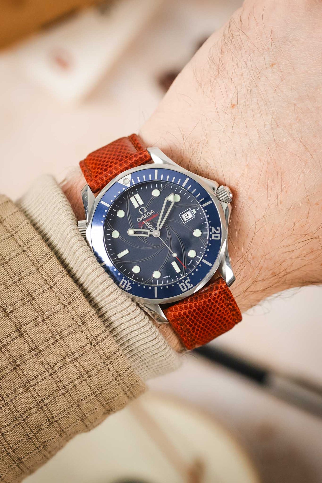 A close-up shot of a wrist wearing a silver Omega Seamaster Diver 300M watch with a blue dial and bezel, paired with a reddish-brown lizard-patterned watch strap. The arm has light skin and is partially covered by a beige, textured sleeve. The background is softly blurred with light, neutral tones.