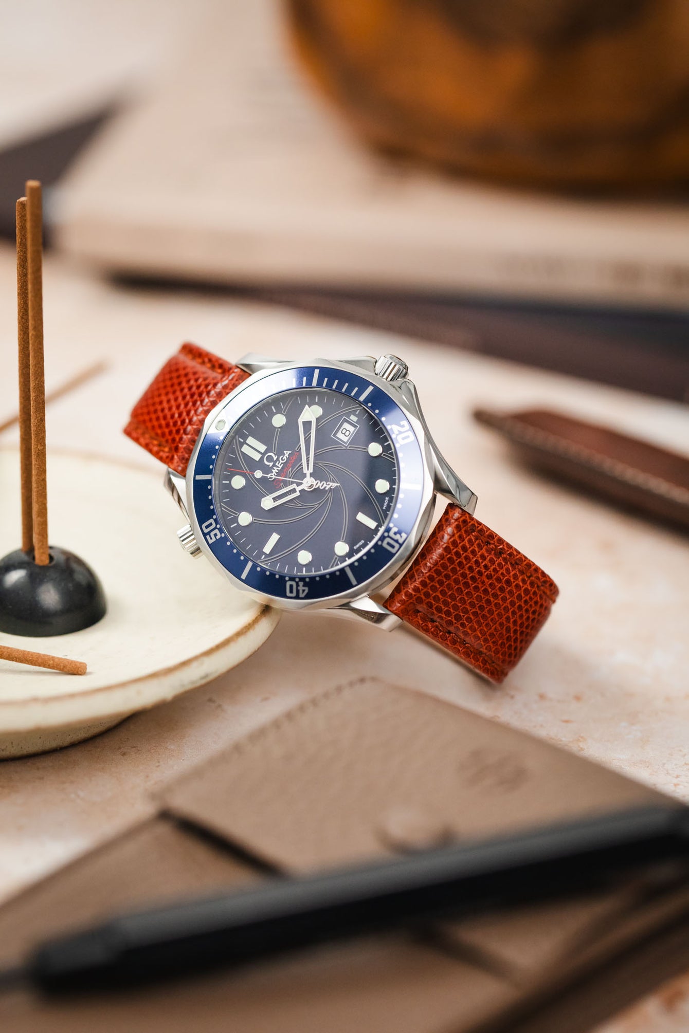 A close-up, eye-level shot of a silver Omega Seamaster Diver 300M watch with a blue dial and bezel, fitted with a reddish-brown lizard-patterned watch strap. The watch is resting on a light, textured surface. In the blurred background, there are incense sticks in a holder, a brown leather item, and a pen.