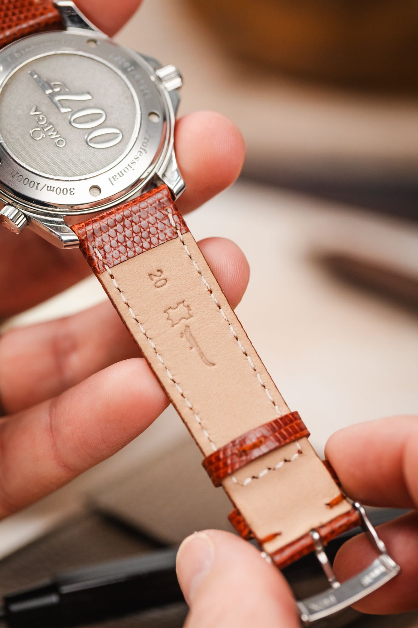 A close-up, overhead shot shows hands holding the back of a silver Omega Seamaster Diver 300M watch. The watch's case back is engraved with "007" and the text "Professional 300m/1000ft". A reddish-brown lizard-patterned watch strap is attached, with its light brown underside visible, bearing a small embossed logo.
