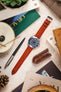 A top-down flat lay composition showcasing a silver Omega Seamaster Diver 300M watch with a blue dial and bezel, fitted with a long, textured reddish-brown watch strap with a silver buckle. The watch is centrally placed and surrounded by various objects on a light, textured surface. These include a black and silver pen, a brown leather tool holder, a magazine, pinecones, and parts of books.