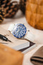 A top-down, slightly angled shot of a silver Tudor Black Bay 58 watch with a blue dial and bezel, fitted with an off-white lizard-patterned strap. The watch is resting on an open book or magazine. A black watch tool is positioned next to it. The background is blurred with warm, earthy tones, showing pinecones and a wooden object.