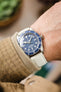 A close-up shot of a wrist wearing a silver Tudor Black Bay 58 watch with a blue dial and bezel, paired with an off-white lizard-patterned watch strap. The arm has light skin and is partially covered by a beige, checkered sleeve. The background is softly blurred with warm, neutral tones.