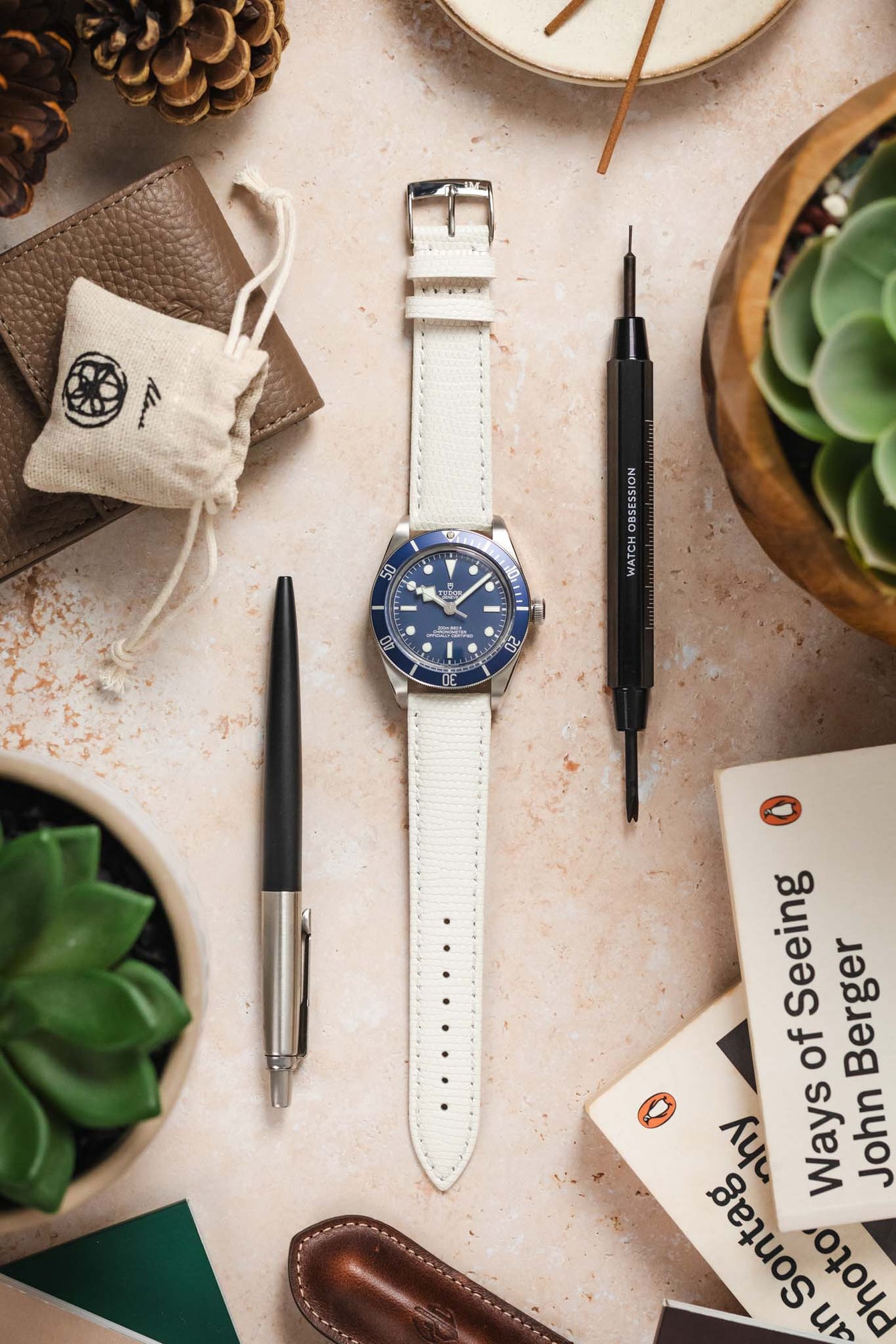 A top-down flat lay composition showcasing a silver Tudor Black Bay 58 watch with a blue dial and bezel, fitted with a long, textured off-white watch strap with a silver buckle. The watch is centrally placed on a light, textured surface. It is surrounded by various objects including pinecones, a brown leather wallet, a small white drawstring bag, a black and silver pen, a black watch tool, a potted succulent plant, and an open book titled "Ways of Seeing".