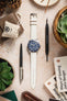 A top-down flat lay composition showcasing a silver Tudor Black Bay 58 watch with a blue dial and bezel, fitted with a long, textured off-white watch strap with a silver buckle. The watch is centrally placed on a light, textured surface. It is surrounded by various objects including pinecones, a brown leather wallet, a small white drawstring bag, a black and silver pen, a black watch tool, a potted succulent plant, and an open book titled "Ways of Seeing".