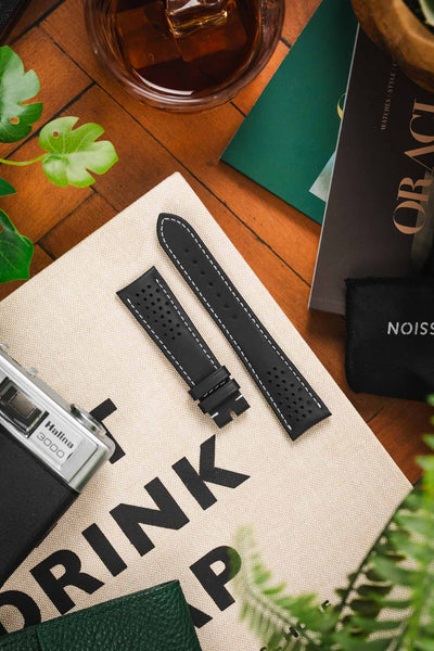 An Omega Watch Strap lay on a cream book in a lifestyle setting. The Strap is black with white stitching and has ventilation holes at either end.