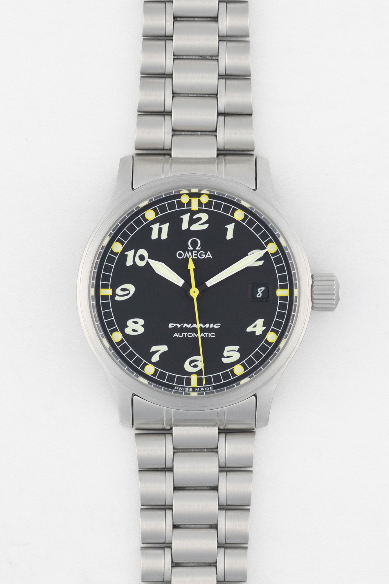 A top-down studio shot of an Omega Dynamic automatic watch with a black dial, white Arabic numerals, and a stainless steel link bracelet. The dial features a yellow center seconds hand and a date window at the 3 o'clock position.