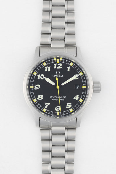 A top-down studio shot of an Omega Dynamic automatic watch with a black dial, white Arabic numerals, and a stainless steel link bracelet. The dial features a yellow center seconds hand and a date window at the 3 o'clock position.