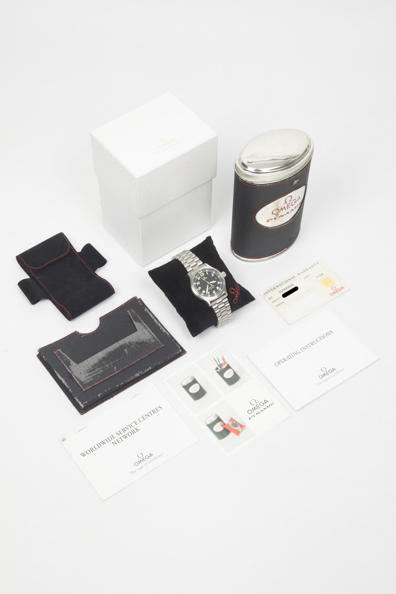 A wide shot of the "full set" including the Omega Dynamic watch on a black cushion, its original white outer box, a unique tin storage case, the international warranty card, and operating instructions manuals.