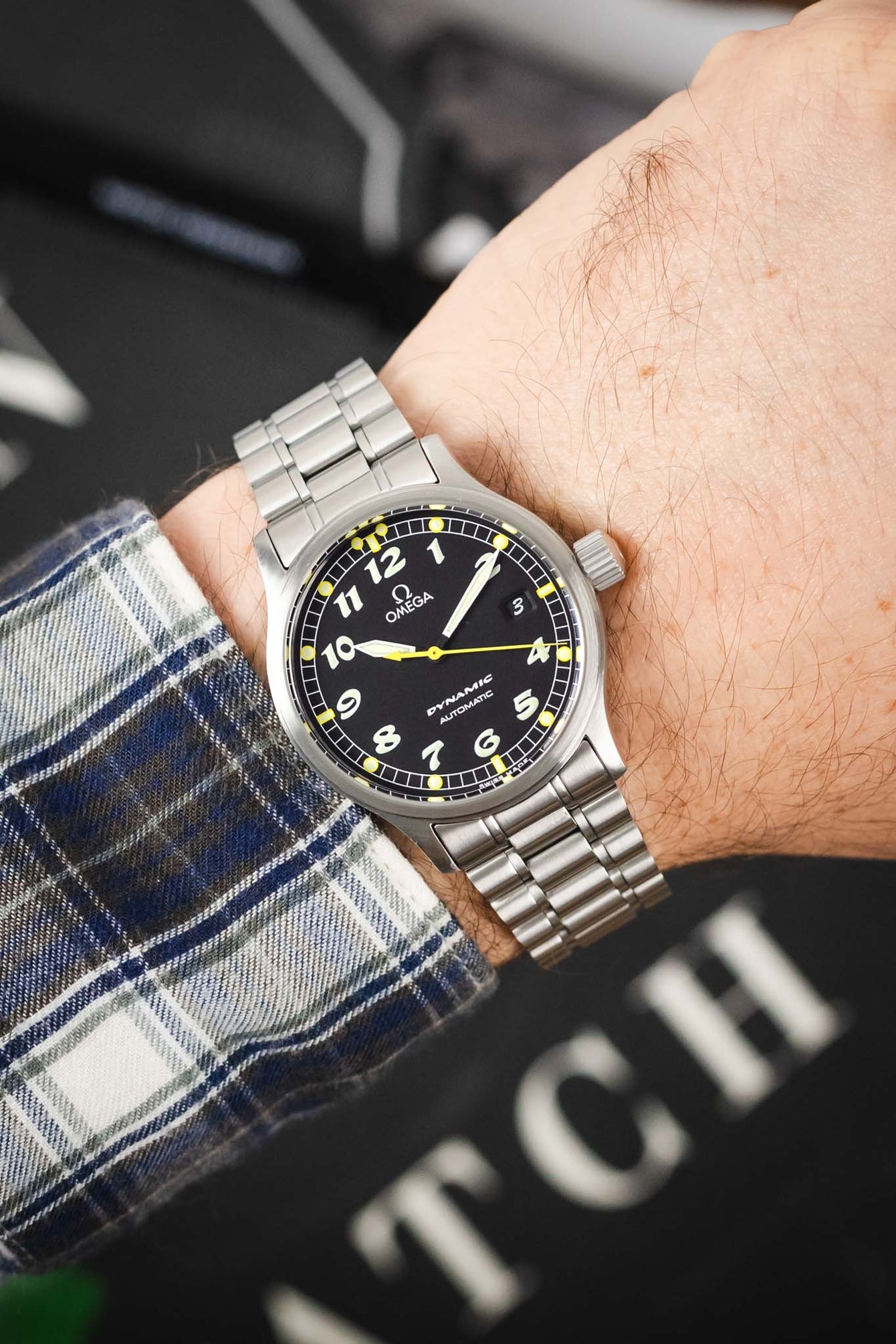 A close-up of the watch being worn on a wrist, paired with a blue and white plaid flannel shirt, showing how the 36mm case sits on the arm.