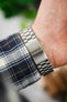 A view of the underside of a wrist showing the brushed stainless steel Omega-branded clasp and the underside of the plaid shirt sleeve.