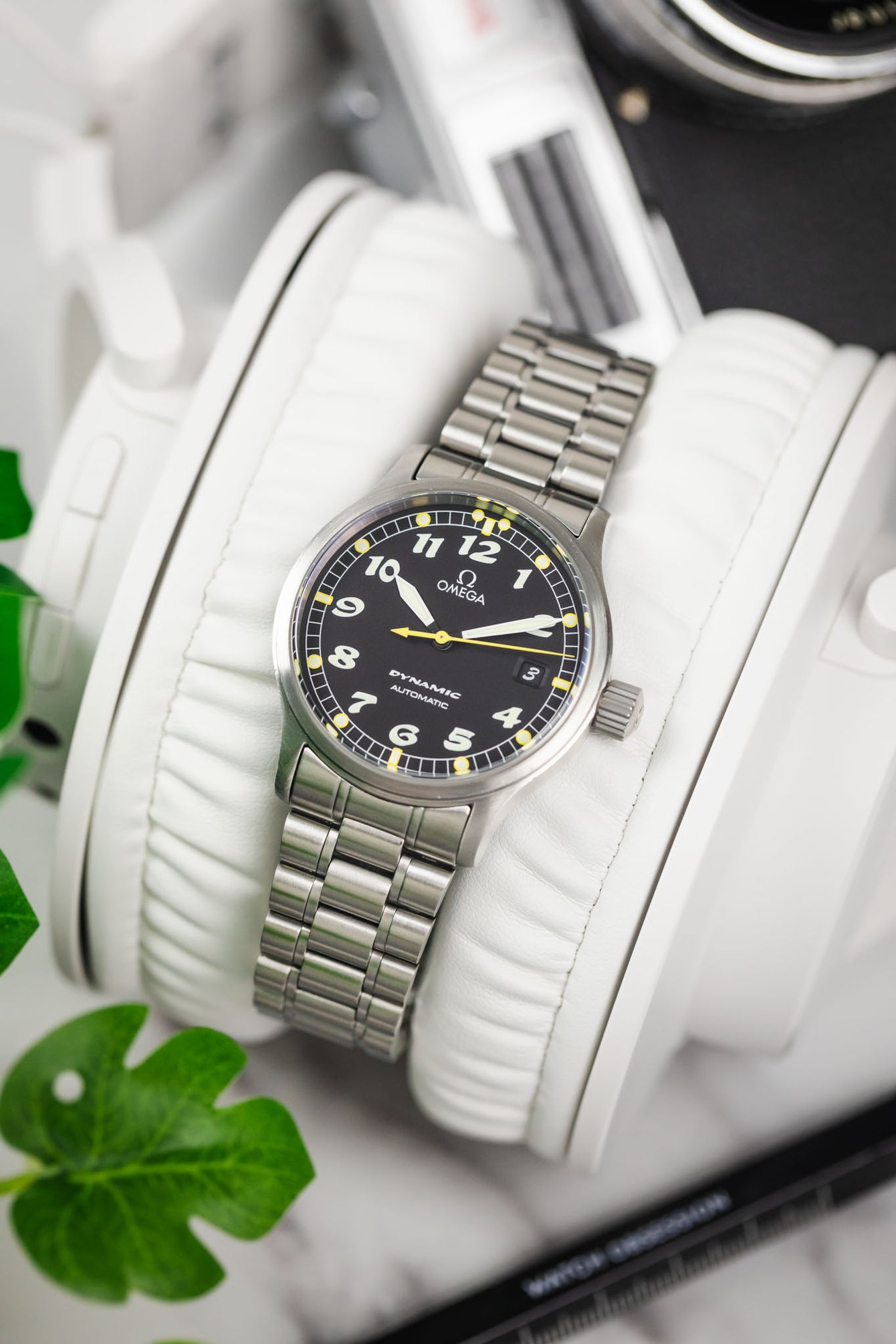 A lifestyle photograph of the Omega Dynamic watch draped over a pair of white leather headphone cushions, with a vintage camera blurred in the background.