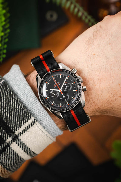 An overhead wrist shot of an Omega Speedy Ultraman, its being worn on a black and orange nylon strap with a grey jumper sleeve and checked jacket.