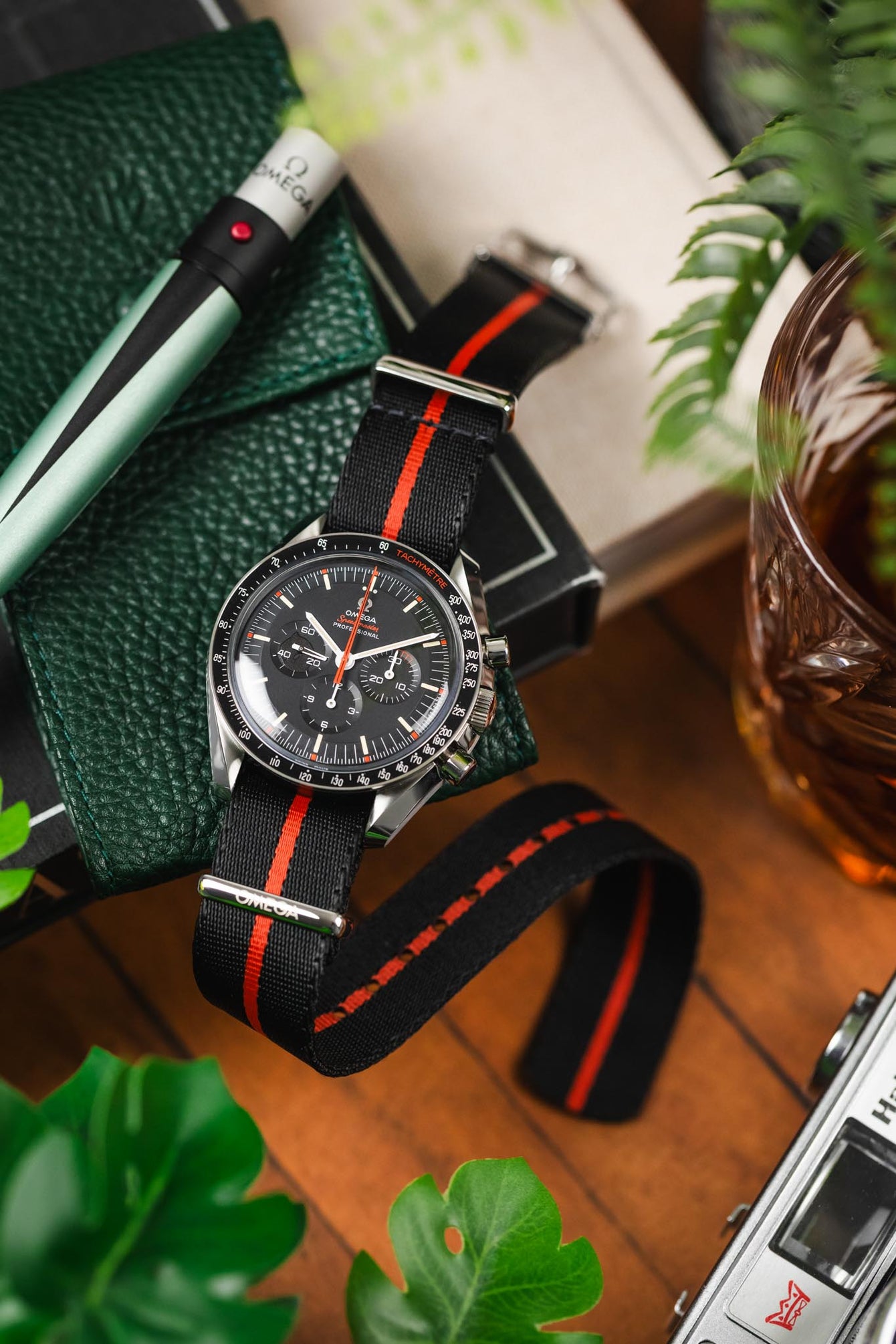 A Speedmaster Ultraman lay on a green leather watch pouch alongside a glass of whiskey, vintage camera and an Omega black light torch.