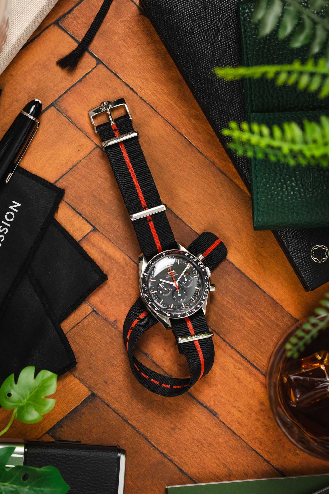 An overhead lay flat shot of an Speedy Tuesday Ultraman with its strap looped through and underneath itself. The watch is placed on a wooden parquet flooring and is surrounded by watch paraphernalia.