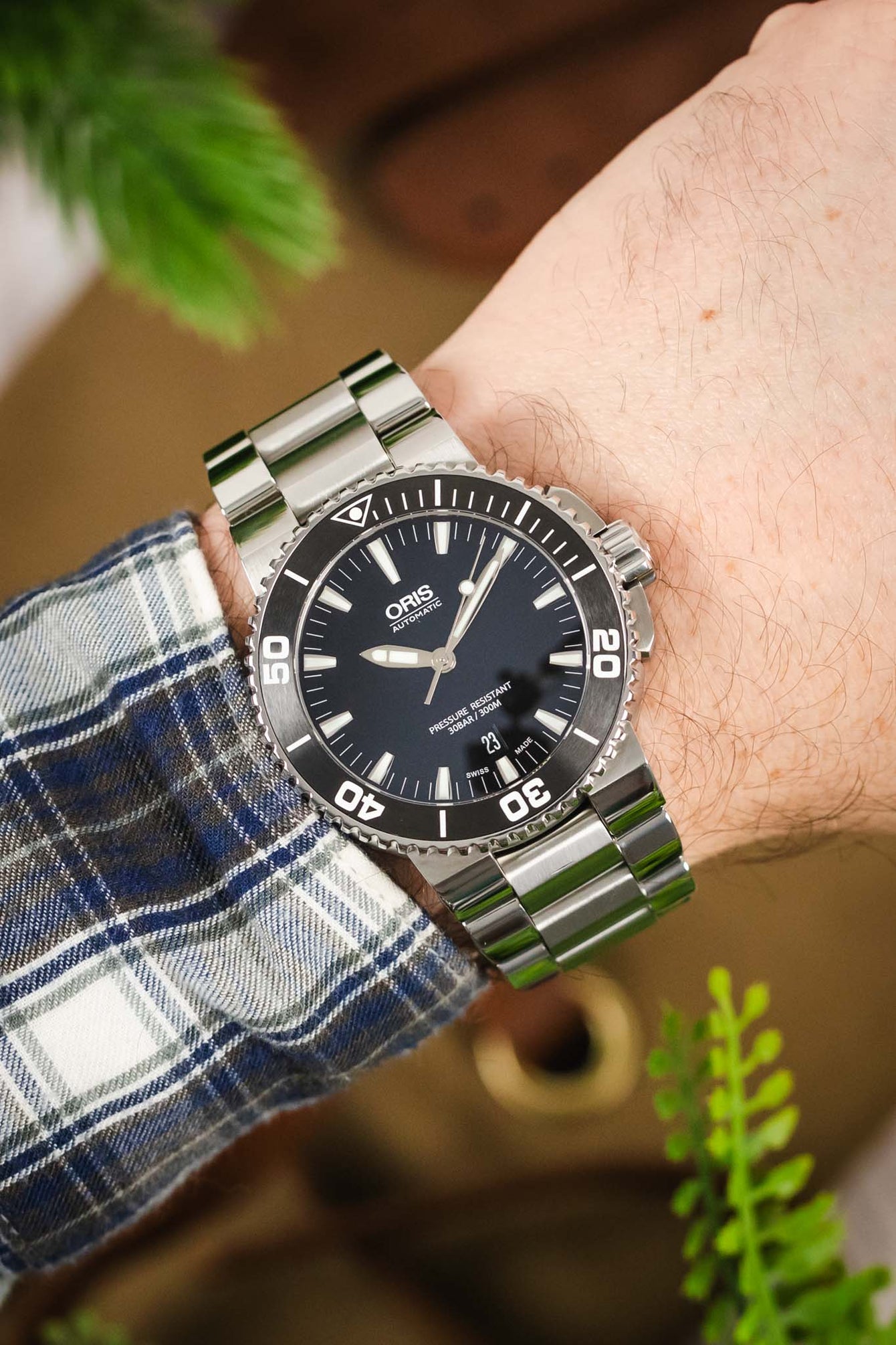 A wrist shot of a person wearing an Oris Aquis Date with a stainless steel bracelet, paired with a blue, grey, and white checkered flannel shirt.