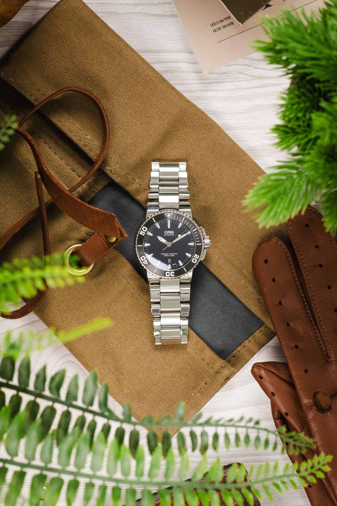 A top-down lifestyle shot featuring an Oris Aquis Date watch on a stainless steel bracelet, centered between a brown leather watch roll, a pair of brown leather gloves, and green foliage.