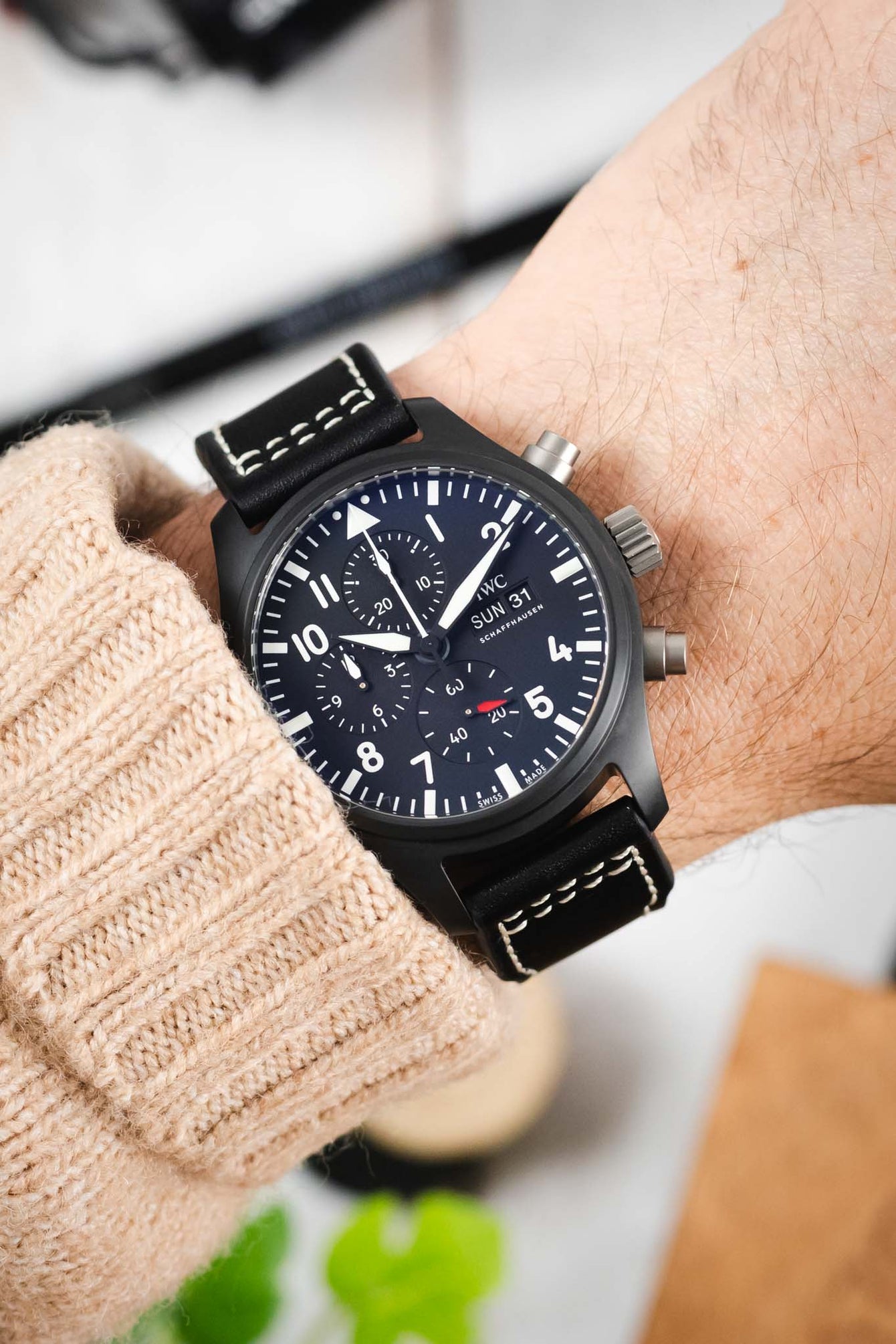Close-up of a black IWC chronograph wristwatch with a matte case, dark dial, bold white numerals, subdials, and a day-date display, worn on a wrist with a black leather strap featuring white contrast stitching, paired with a beige knit sleeve against a softly blurred indoor background.