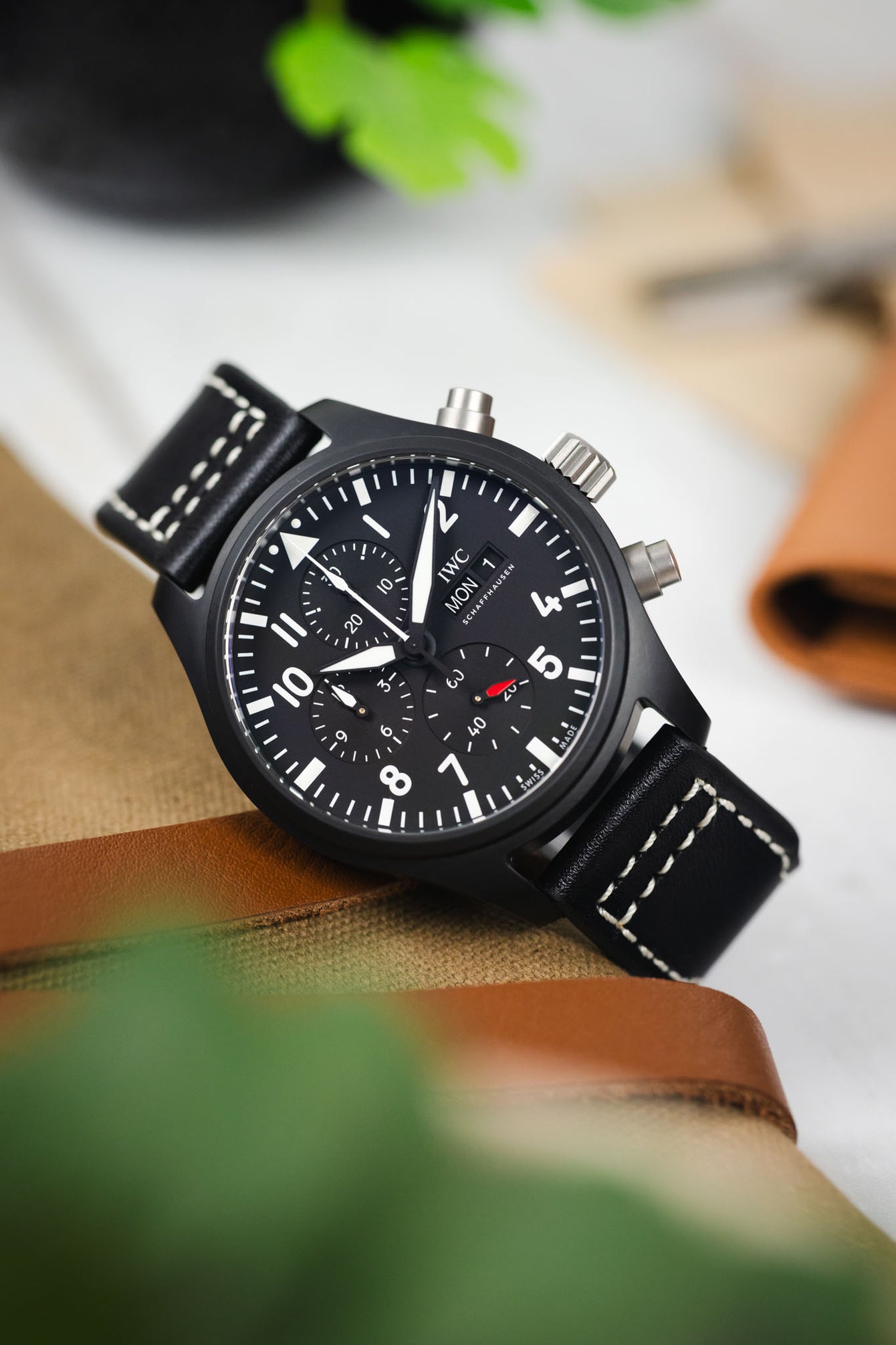 Close-up of a black IWC chronograph wristwatch with a matte black case, dark dial, bold white numerals, three subdials, and a day-date display, paired with a black leather strap featuring white contrast stitching. The watch is angled on a tan canvas surface with a brown leather strap beneath it, while blurred green leaves and workshop items appear in the softly lit background.