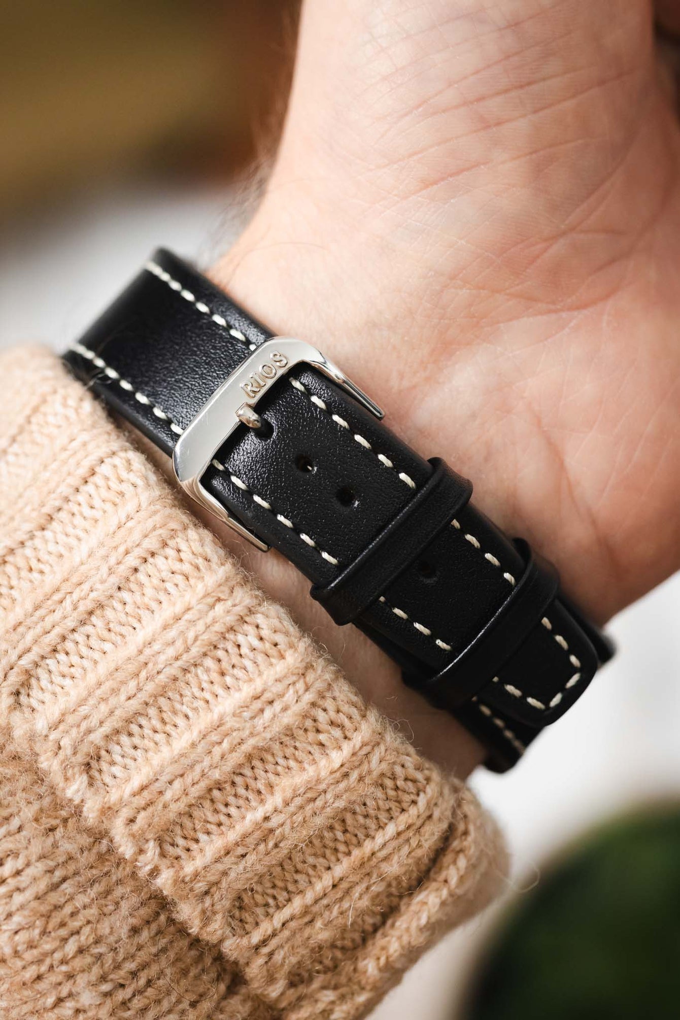 Macro close-up of a black leather watch strap with white contrast stitching fastened on a wrist, showing a silver buckle engraved with “RIOS,” paired with a beige knit sleeve and set against a softly blurred background with warm tones and greenery.
