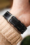 Macro close-up of a black leather watch strap with white contrast stitching fastened on a wrist, showing a silver buckle engraved with “RIOS,” paired with a beige knit sleeve and set against a softly blurred background with warm tones and greenery.