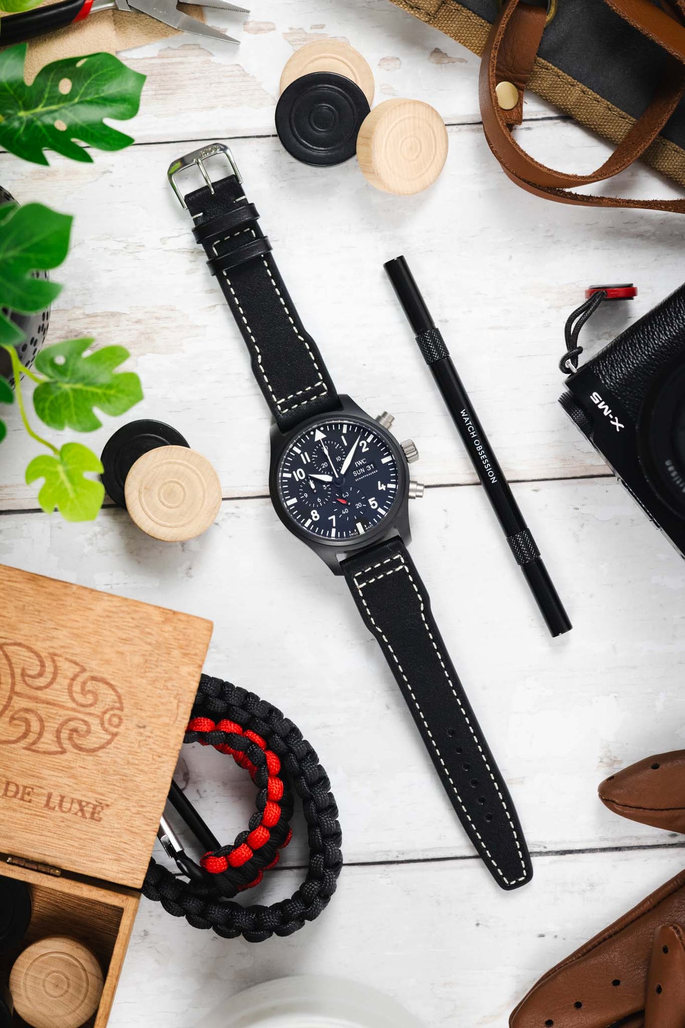 Flat lay of a black IWC chronograph wristwatch with a matte case, dark dial, bold white numerals, three subdials, and a day-date display, paired with a black leather strap with white contrast stitching. The watch is centered on a light wooden surface, surrounded by a pen labeled “Watch Obsession,” a camera, paracord bracelet, wooden game pieces, leather accessories, small tools, and green plant leaves.