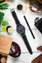 Flat lay of a black IWC chronograph wristwatch with a matte case, dark dial, bold white numerals, three subdials, and a day-date display, paired with a black leather strap with white contrast stitching. The watch is centered on a light wooden surface, surrounded by a pen labeled “Watch Obsession,” a camera, paracord bracelet, wooden game pieces, leather accessories, small tools, and green plant leaves.
