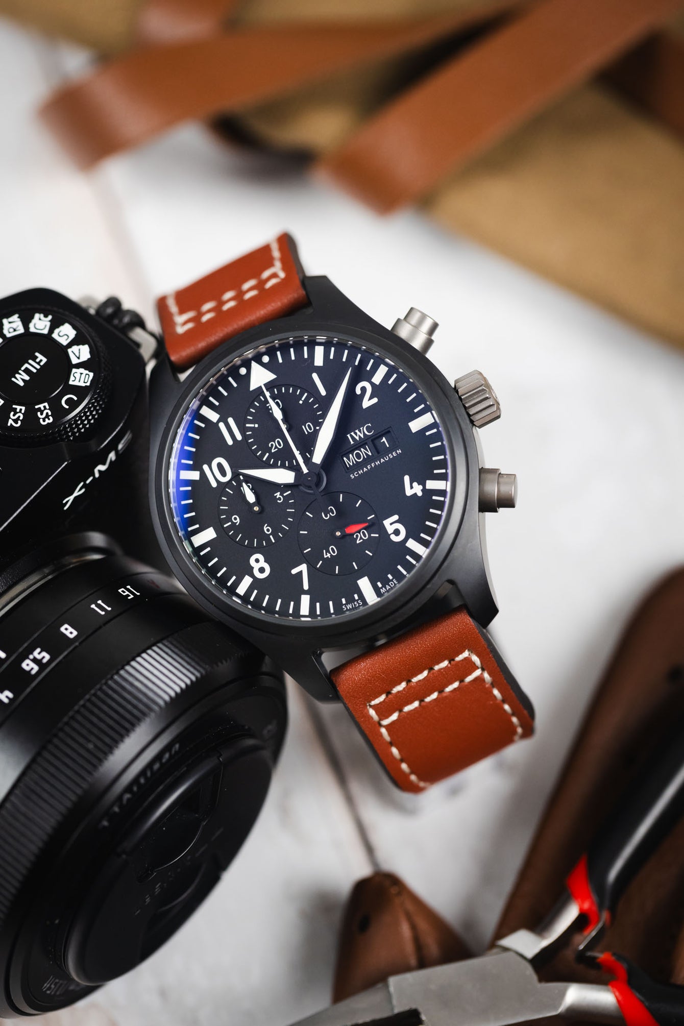 Close-up of a black IWC chronograph wristwatch with a matte case, dark dial, bold white numerals, three subdials, and a day-date display, paired with a cognac-brown leather strap with white contrast stitching. The watch is positioned next to a black camera body and lens on a light wooden surface, with tools and leather accessories softly blurred in the background.