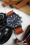 Close-up of a black IWC chronograph wristwatch with a matte case, dark dial, bold white numerals, three subdials, and a day-date display, paired with a cognac-brown leather strap with white contrast stitching. The watch is positioned next to a black camera body and lens on a light wooden surface, with tools and leather accessories softly blurred in the background.