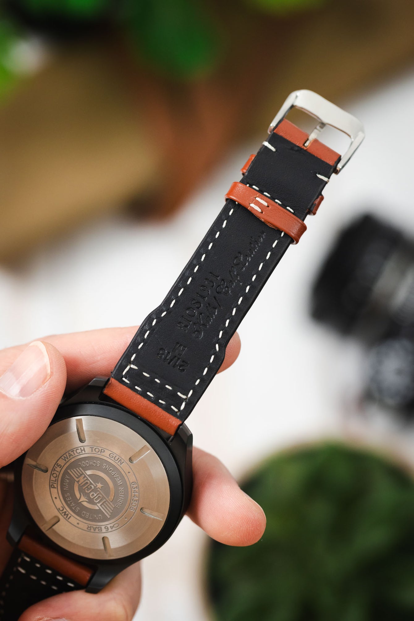 Macro close-up of the back of a black IWC chronograph wristwatch held in hand, showing the engraved caseback with markings including “IWC Pilot’s Watch Top Gun,” along with a black leather strap lined with cognac-brown leather accents and white contrast stitching. The strap interior features embossed branding text, and the silver buckle is visible at the top, set against a softly blurred background with warm tones, greenery, and workshop elements.