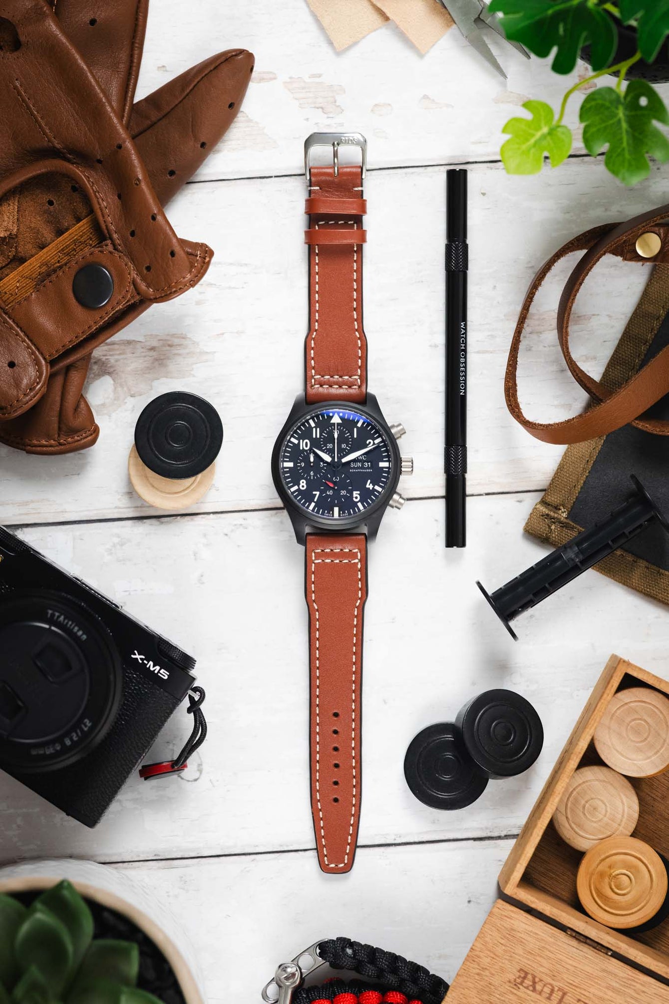 Flat lay of a black IWC chronograph watch with a cognac leather strap, surrounded by a camera, gloves, tools, and wooden game pieces on a white surface.