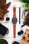 Flat lay of a black IWC chronograph watch with a cognac leather strap, surrounded by a camera, gloves, tools, and wooden game pieces on a white surface.