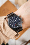 Close-up of a black IWC chronograph watch with a dark brown leather strap worn on a wrist, paired with a beige knit sleeve.