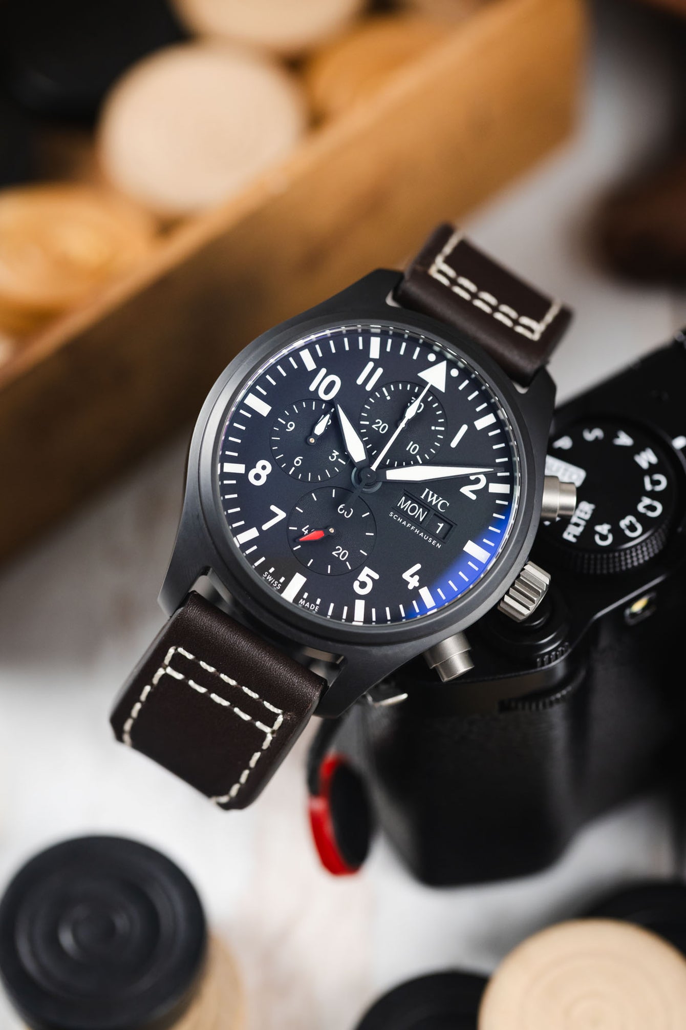 Close-up of a black IWC chronograph watch with a dark brown leather strap resting against a camera on a light surface.