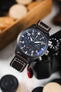 Close-up of a black IWC chronograph watch with a dark brown leather strap resting against a camera on a light surface.