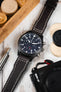 Close-up of a black IWC chronograph watch with a dark brown leather strap lying on a light surface beside a camera and wooden game pieces.