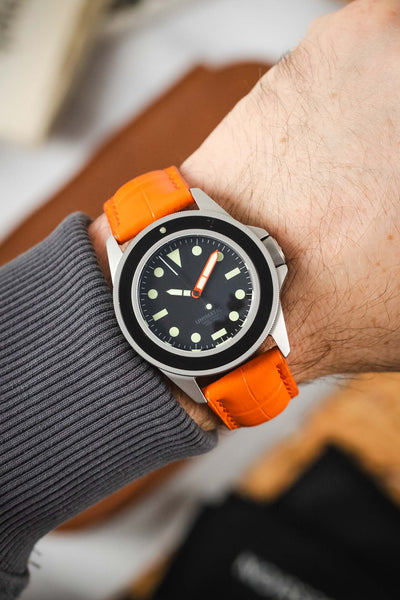 
A close-up shot of a person's wrist wearing a UNIMATIC watch with a black dial and a vibrant orange crocodile-embossed leather watch strap. The person is wearing a gray long-sleeved shirt. The background is softly blurred.