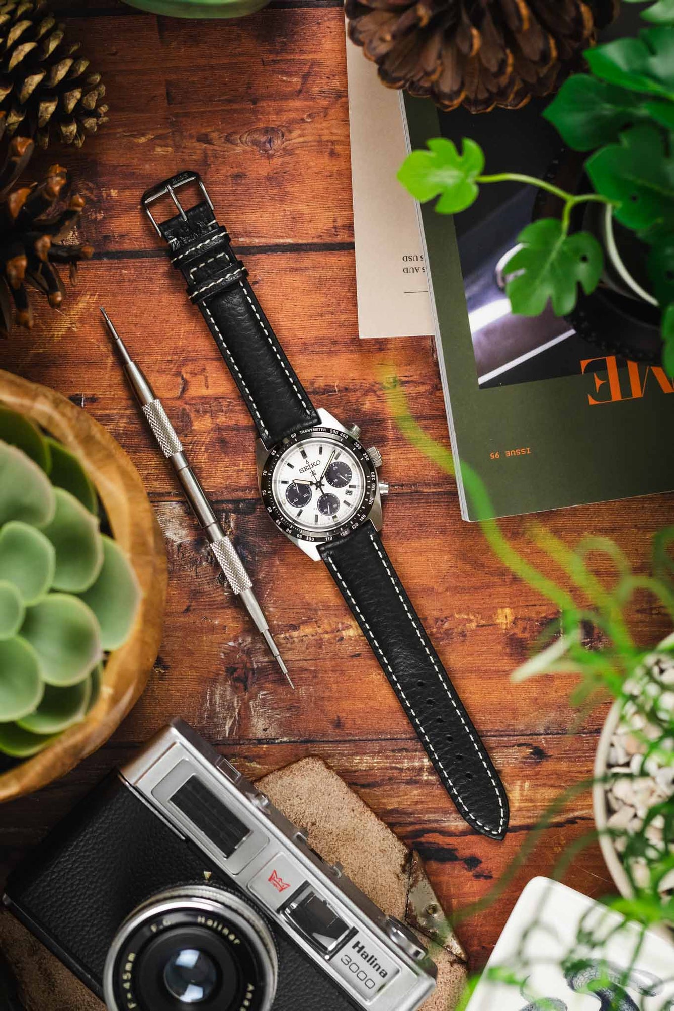A Seiko chronograph watch with a black RIOS1931 Montana leather strap with white stitching, displayed on a wooden table with a camera, plants, and a watchmaker's tool.