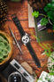 A Seiko chronograph watch with a black RIOS1931 Montana leather strap with white stitching, displayed on a wooden table with a camera, plants, and a watchmaker's tool.