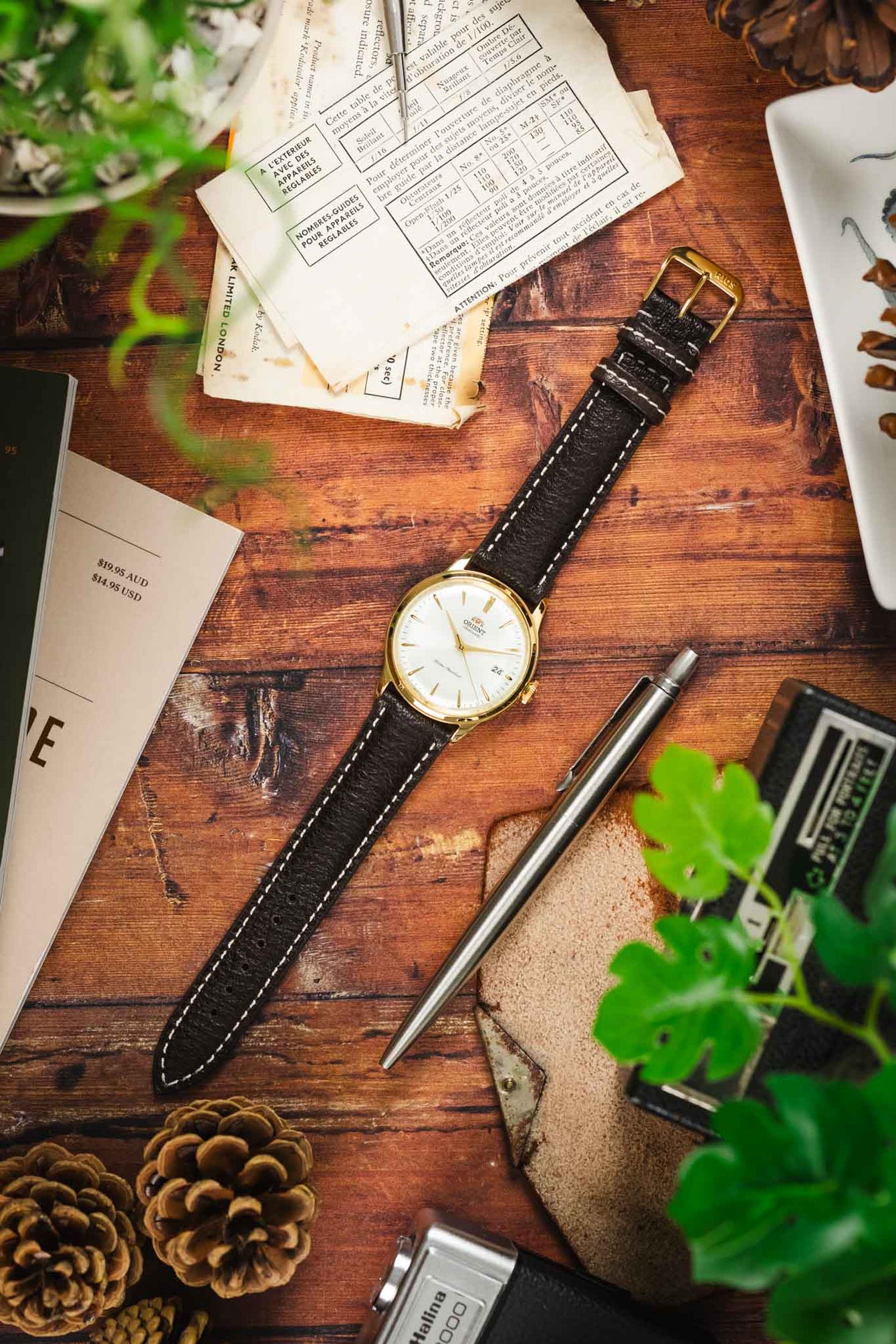 An Orient watch with a dark brown RIOS1931 Montana leather strap with white stitching and a gold buckle, on a wooden table with vintage items.
