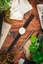 An Orient watch with a dark brown RIOS1931 Montana leather strap with white stitching and a gold buckle, on a wooden table with vintage items.