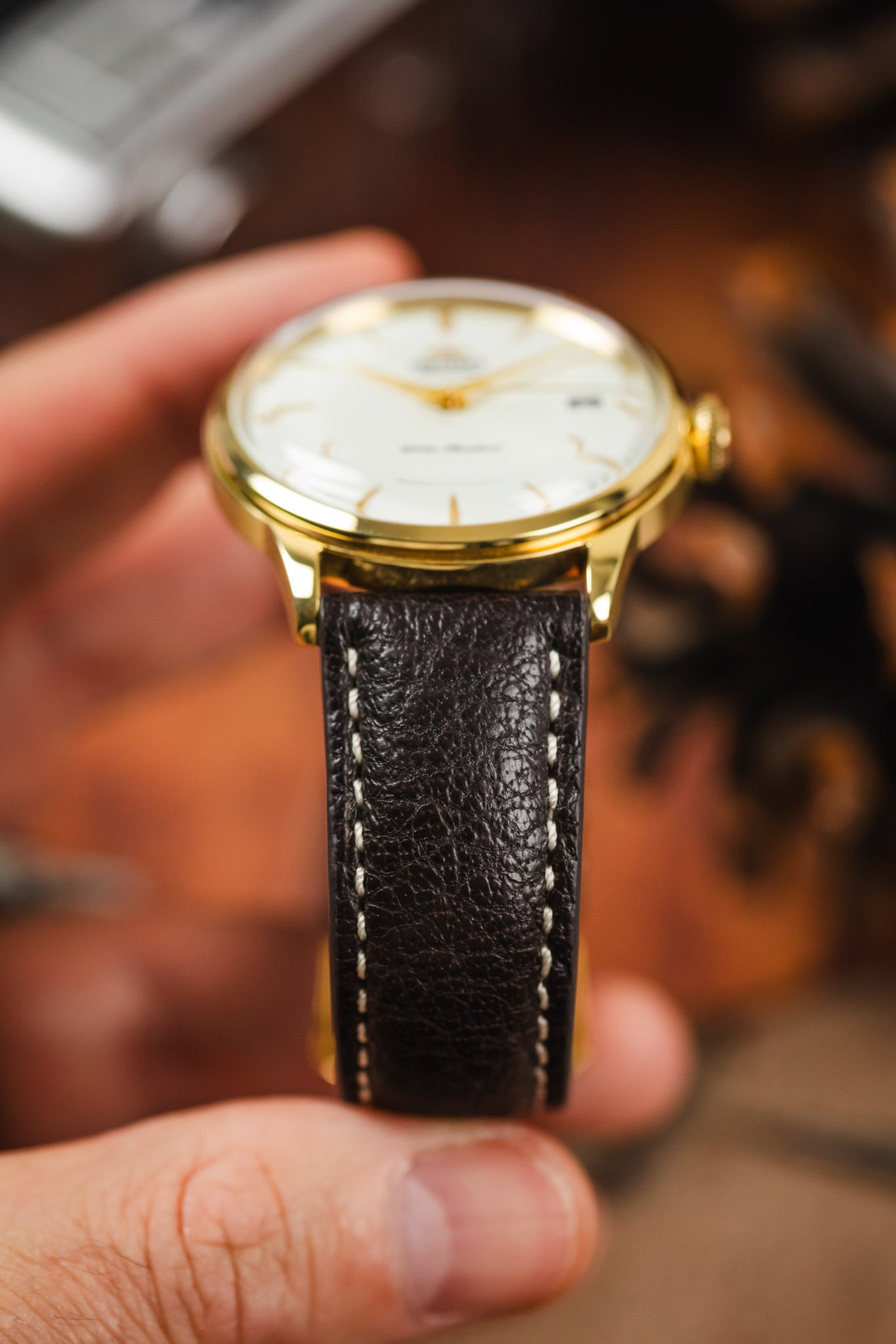 A hand holding an Orient watch on a dark brown RIOS1931 Montana leather strap with white stitching.