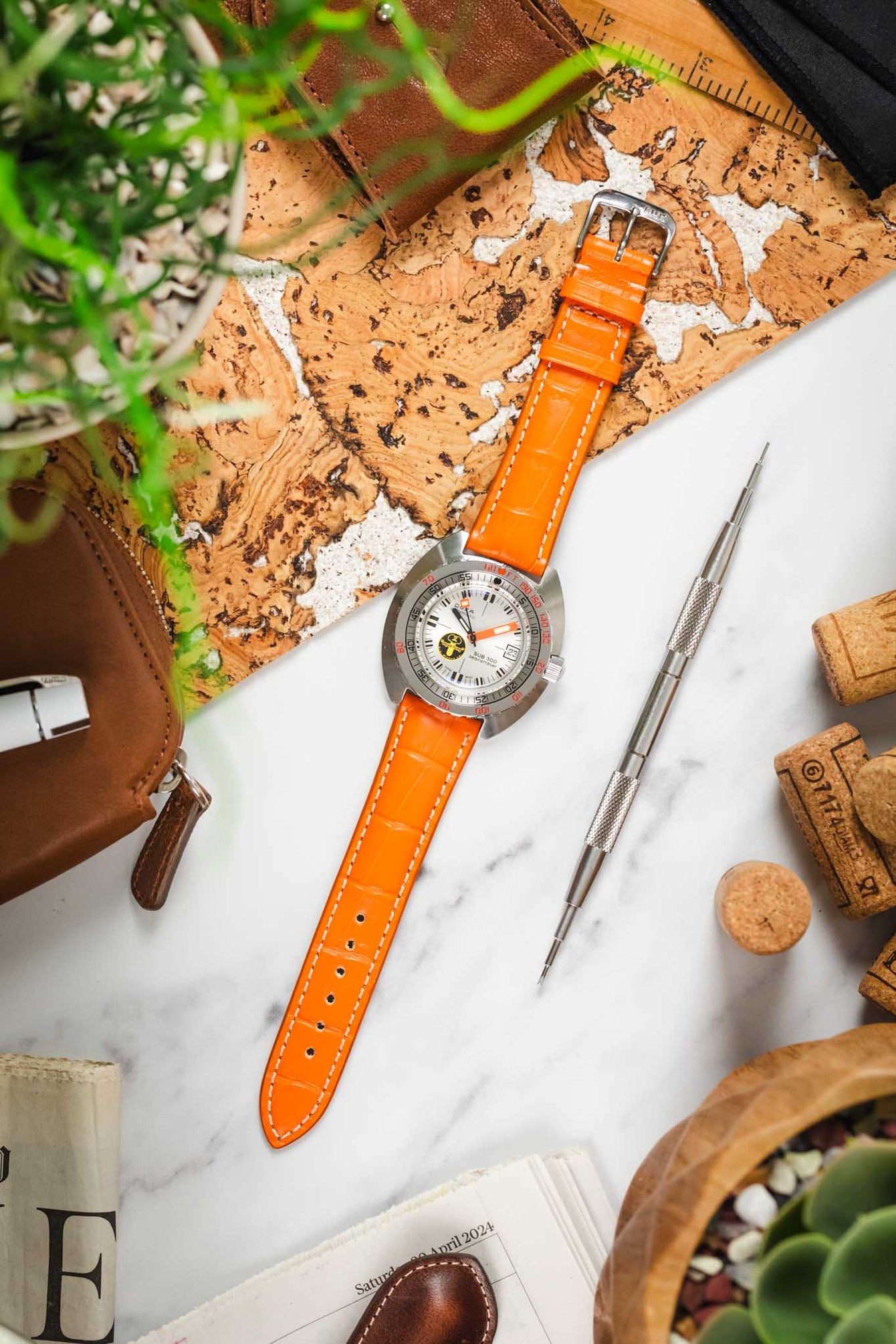 A top-down flat lay shows a DOXA SUB 300 watch with a silver dial and an orange crocodile-embossed leather strap. The watch is positioned on a light surface with a piece of patterned cork. Surrounding the watch are a silver watch tool, corks, a potted succulent, and various other watch-related and decorative elements.